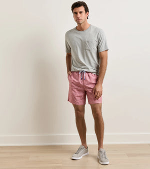 A man stands indoors in front of a plain beige wall on a light wood floor, wearing a grey t-shirt, grey sneakers, and Peter Millar Tails of Summer Swim Trunk featuring pink patterned fabric and an elastic drawstring waistband.