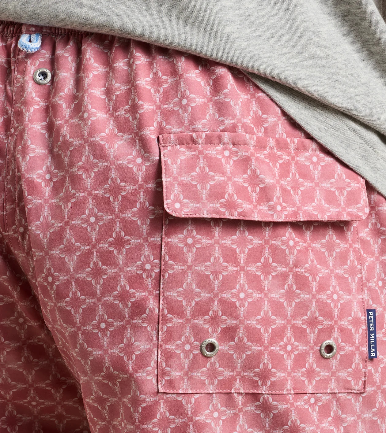 Close-up of a person wearing the Peter Millar Tails of Summer Swim Trunk, featuring a pink patterned design with a flap pocket and visible brand tag, an elastic drawstring waistband, paired with a light gray shirt.