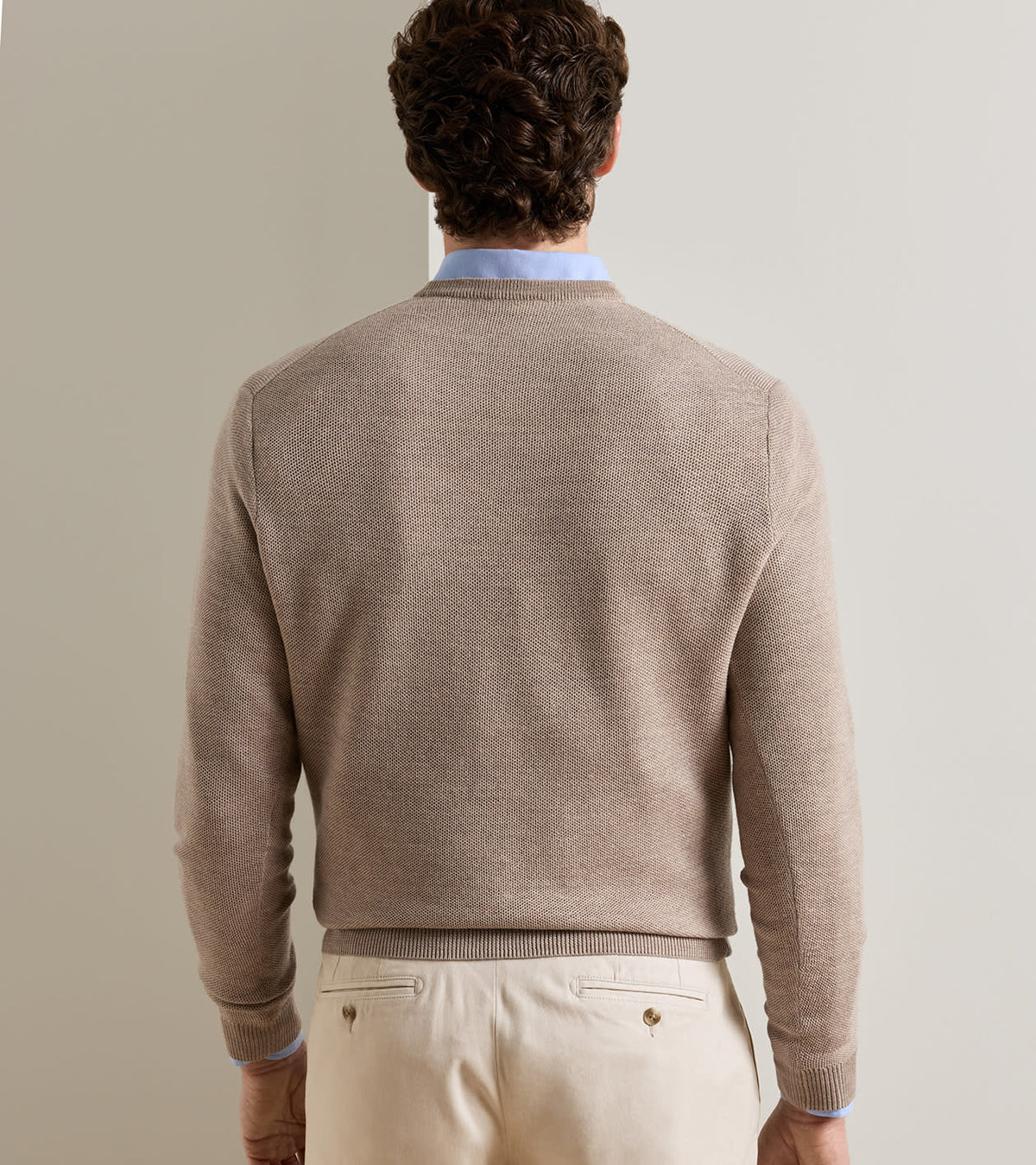 A person seen from behind wears a tan Peter Millar Autumn Crest Honeycomb Crewneck Sweater over a light blue collared shirt and cream trousers.