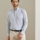 A man is wearing the Peter Millar Bruce Crown Lite Cotton shirt in light blue checkered twill, crafted from easy-care fabric, paired with beige pants and a black belt, standing against a neutral-colored wall.
