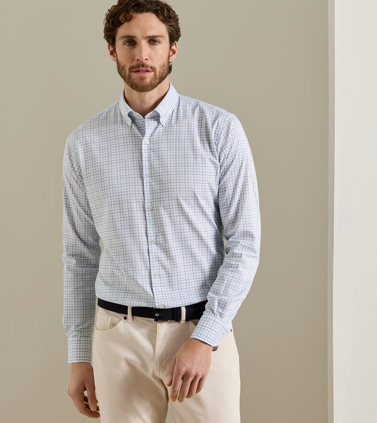 A man is wearing the Peter Millar Bruce Crown Lite Cotton shirt in light blue checkered twill, crafted from easy-care fabric, paired with beige pants and a black belt, standing against a neutral-colored wall.