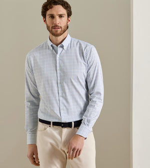 A man is wearing the Peter Millar Bruce Crown Lite Cotton shirt in light blue checkered twill, crafted from easy-care fabric, paired with beige pants and a black belt, standing against a neutral-colored wall.