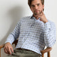 A man with light brown hair, wearing the Peter Millar Lucas Performance Twill Sport Shirt in a blue and white plaid pattern and olive green pants, sits on a wooden chair, resting his head on his hand while looking at the camera.
