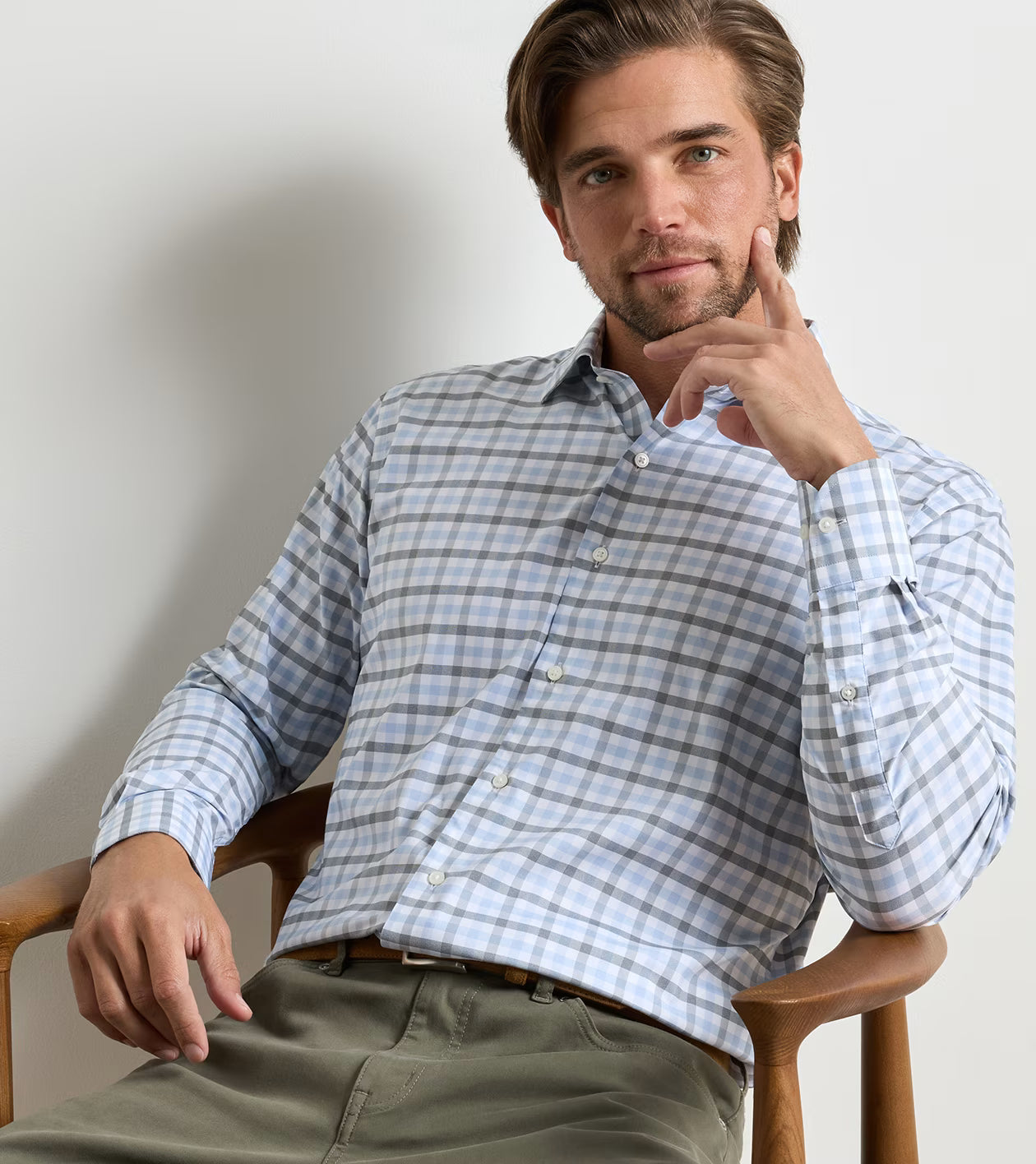 A man with light brown hair, wearing the Peter Millar Lucas Performance Twill Sport Shirt in a blue and white plaid pattern and olive green pants, sits on a wooden chair, resting his head on his hand while looking at the camera.