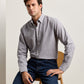A man with short brown hair, dressed in the Peter Millar Devonport Chambray Cotton-Stretch Sport Shirt in light grey and blue jeans for a casual look, sits on a wooden chair against a plain background.