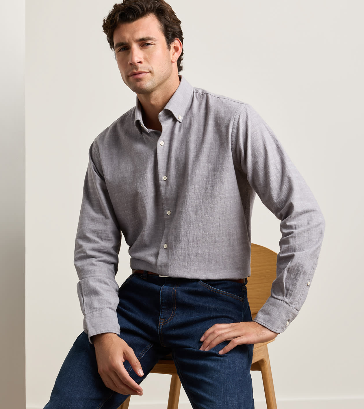A man with short brown hair, dressed in the Peter Millar Devonport Chambray Cotton-Stretch Sport Shirt in light grey and blue jeans for a casual look, sits on a wooden chair against a plain background.