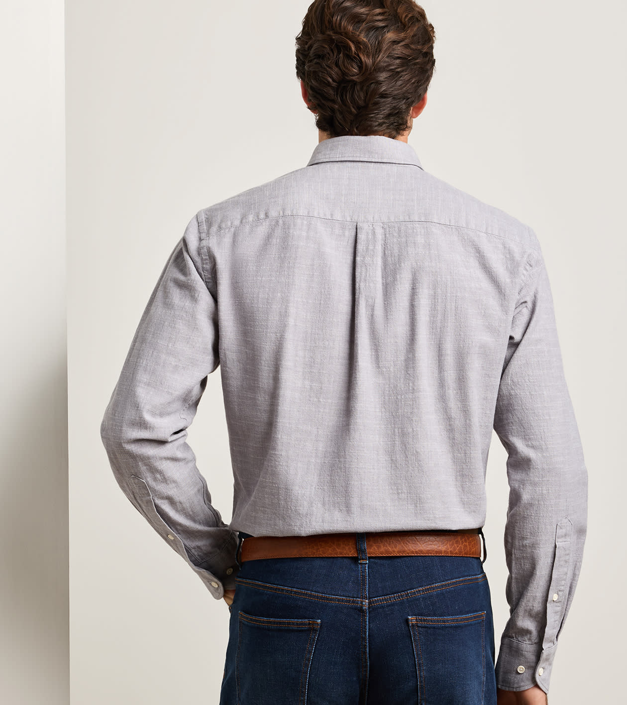 A man stands with his back to the camera, wearing the Peter Millar Devonport Chambray Cotton-Stretch Sport Shirt in light gray, tucked into blue jeans and paired with a brown belt for a relaxed casual style.