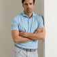 A man in the Peter Millar Keynote Performance Jersey Polo, featuring light blue stripes and four-way stretch, paired with light gray pants, stands with arms crossed, leaning against a white wall.
