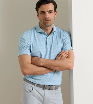 A man in the Peter Millar Keynote Performance Jersey Polo, featuring light blue stripes and four-way stretch, paired with light gray pants, stands with arms crossed, leaning against a white wall.
