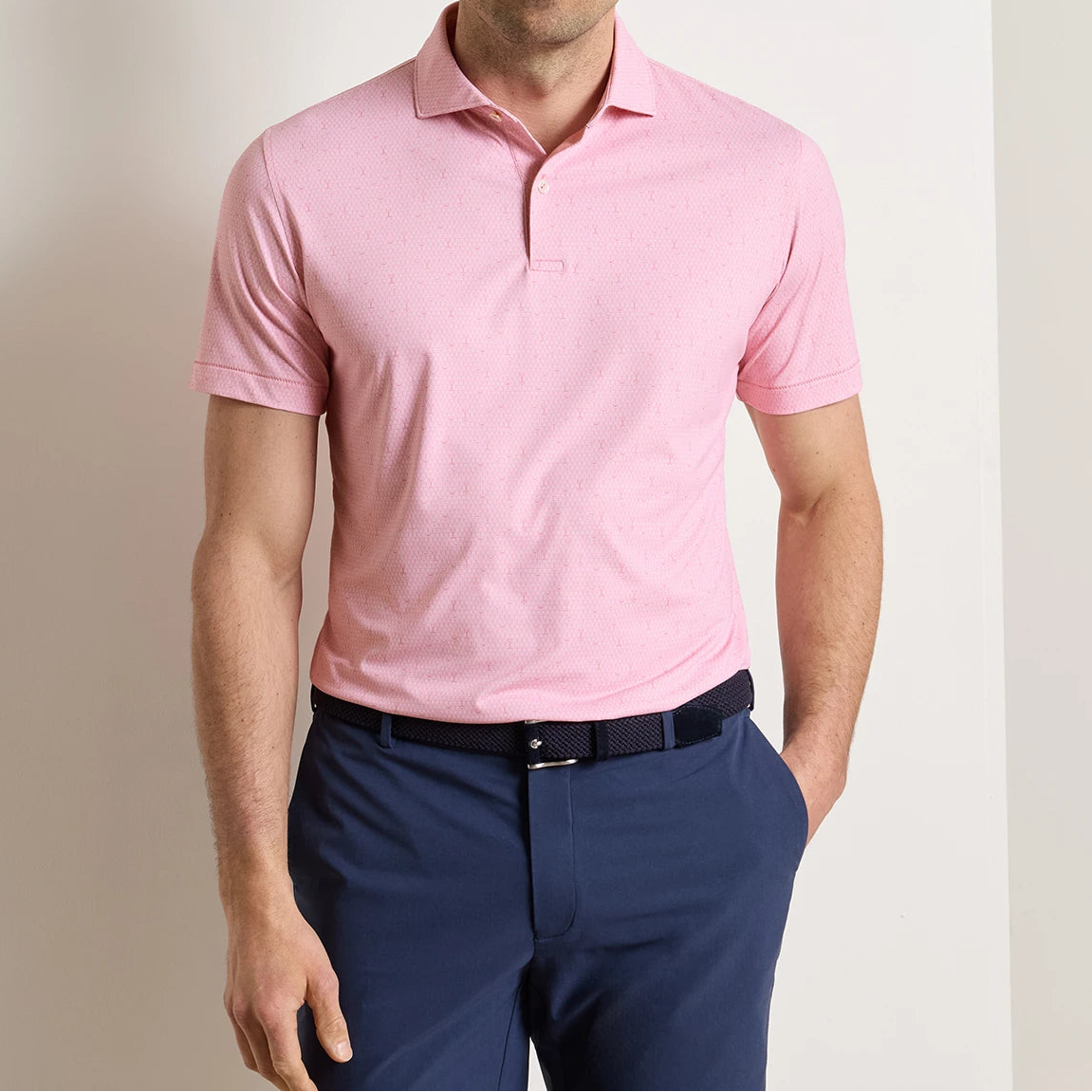 A man wearing the Peter Millar Fifty-Fifty Performance Jersey Polo in light pink with four-way stretch, paired with navy blue pants, stands against a plain white wall.