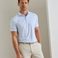A man stands indoors wearing the Peter Millar Gingko Performance Jersey Polo in light blue, paired with beige pants and a belt, looking at the camera with a neutral expression.
