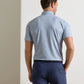 A man stands with his back to the camera, wearing the Peter Millar Limoncello Performance Jersey Polo—a light blue patterned short-sleeve shirt featuring UPF 50+ sun protection—tucked into navy pants with a dark belt.
