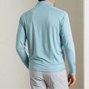 A man is shown from the back, indoors near a white wall, wearing the Peter Millar Stealth Performance Quarter-Zip in light blue with long sleeves and light gray pants.