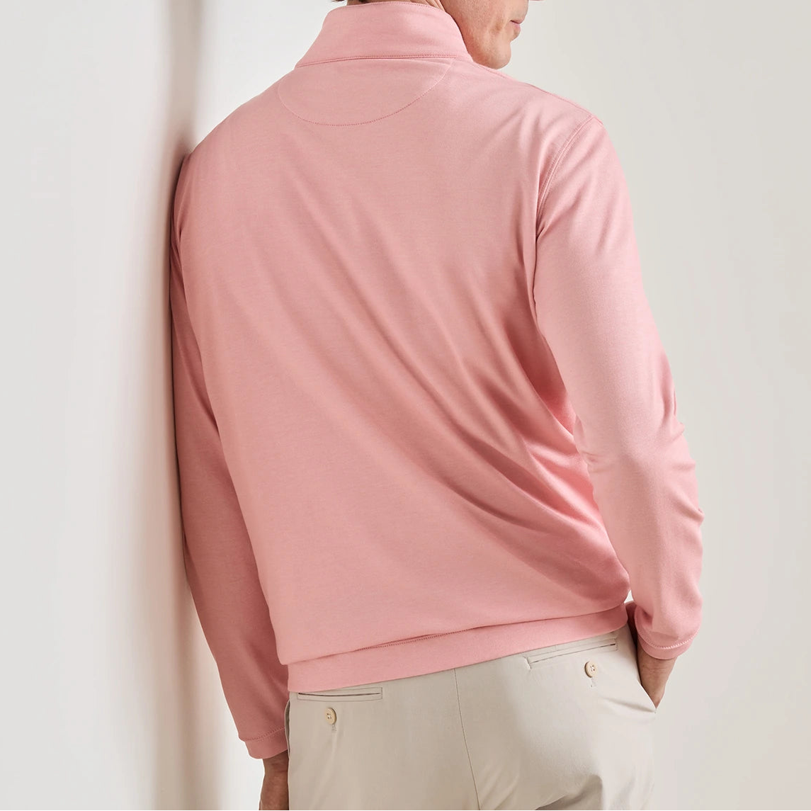 A person is seen from behind, leaning against a white wall, wearing the Peter Millar Albatross Cotton-Blend Pique Quarter-Zip in light pink and a pair of light beige pants.