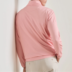 A person is seen from behind, leaning against a white wall, wearing the Peter Millar Albatross Cotton-Blend Pique Quarter-Zip in light pink and a pair of light beige pants.