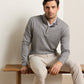 A man is wearing the Peter Millar Grant Knit Quarter-Zip sweater in grey paired with beige pants, seated on a wooden bench against a plain, light-colored background.