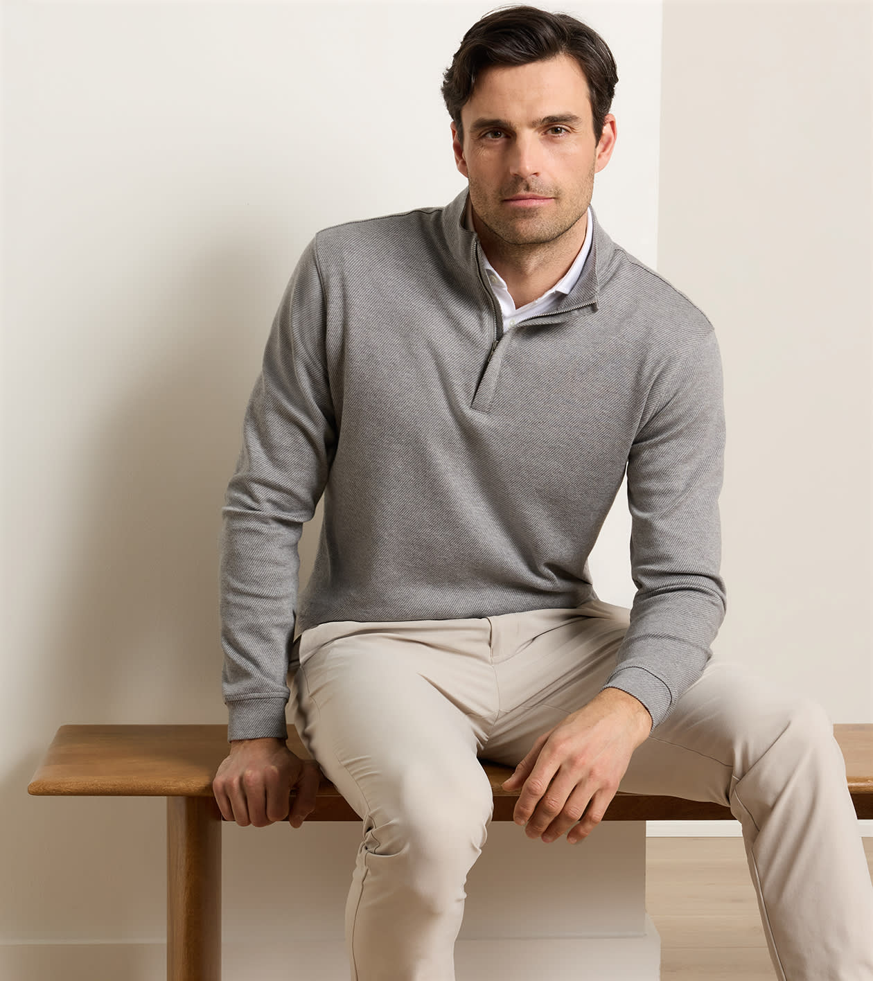 A man is wearing the Peter Millar Grant Knit Quarter-Zip sweater in grey paired with beige pants, seated on a wooden bench against a plain, light-colored background.
