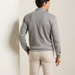 A man stands indoors facing away, wearing the Peter Millar Grant Knit Quarter-Zip in gray—a long-sleeve shirt crafted from a soft cotton-cashmere blend—paired with light beige pants, his left arm slightly bent.