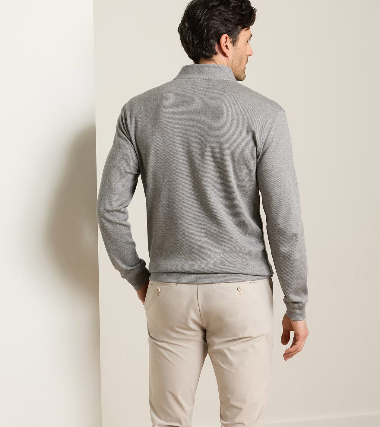 A man stands indoors facing away, wearing the Peter Millar Grant Knit Quarter-Zip in gray—a long-sleeve shirt crafted from a soft cotton-cashmere blend—paired with light beige pants, his left arm slightly bent.