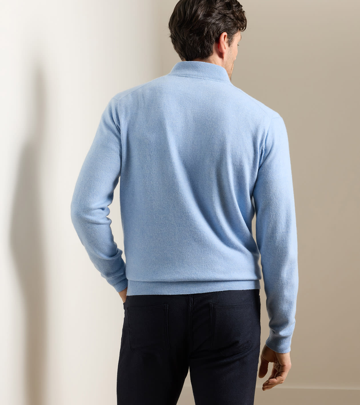 A man stands with his back to the camera, wearing the Peter Millar Artisan Crafted Cashmere Flex Quarter-Zip in light blue and dark pants, in a softly lit indoor setting.