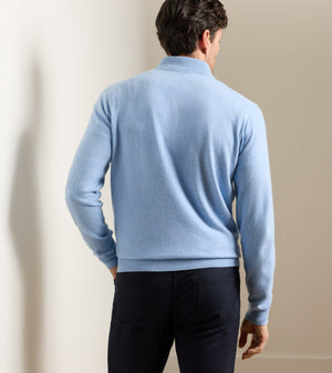 A man stands with his back to the camera, wearing the Peter Millar Artisan Crafted Cashmere Flex Quarter-Zip in light blue and dark pants, in a softly lit indoor setting.