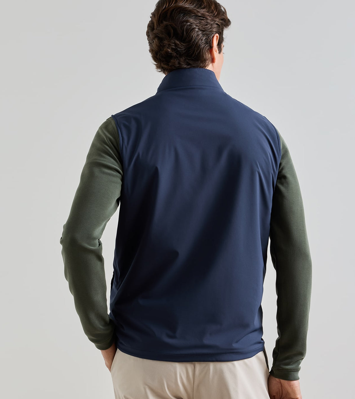 View from behind of a person wearing a navy blue water-resistant Peter Millar Ryder Reversible Vest over an olive green long-sleeved shirt and light-colored trousers.