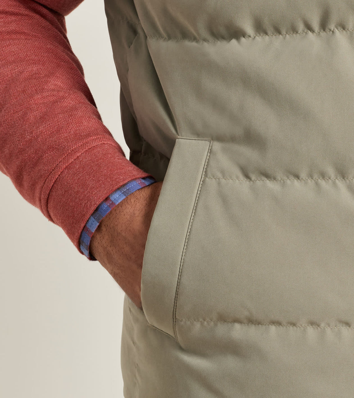 Close-up of a person wearing a light olive Bedford Quilted Vest by Peter Millar over a red long-sleeved shirt, with a hand tucked into the pocket.