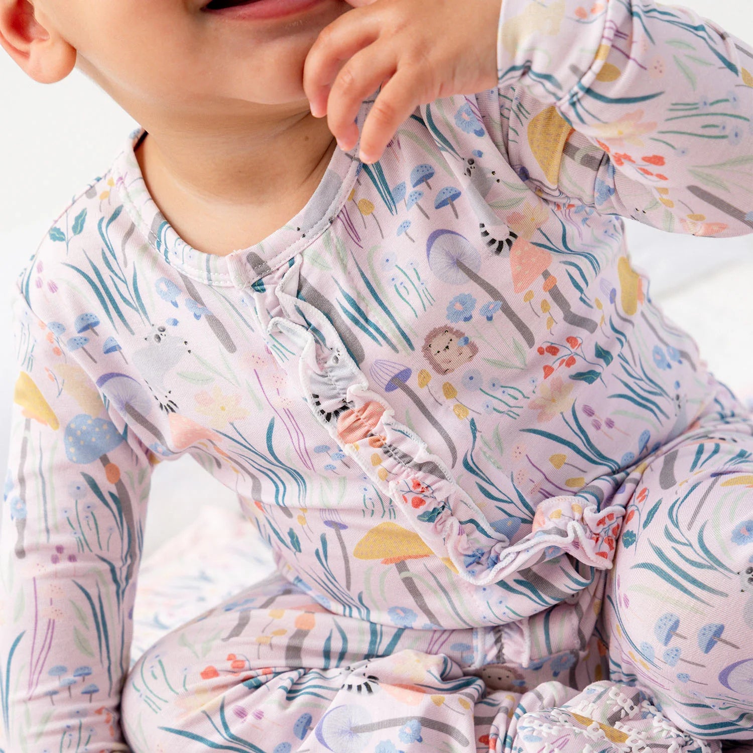 A baby girl in the Magnetic Me Shroom and Bloom with Ruffle Modal Magnetic Footie featuring a whimsical forest print sits on a light surface, smiling and touching her face.