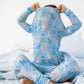 A child on a bed with light blue striped sheets pulls a Magnetic Me Baby and Kids' Party Animals Glow in the Dark Modal Magnetic PJ Set over their head, making dressing easy.