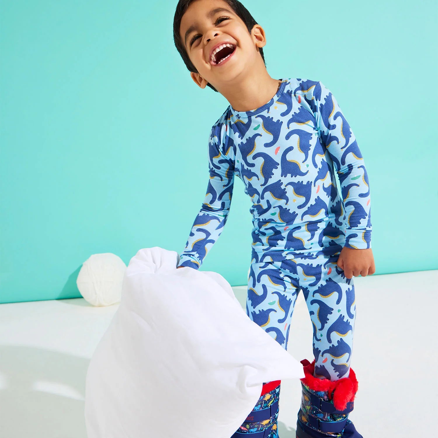 A young boy in Magnetic Me's Baby Boys' and Boys' Necks Generation Modal PJ Set stands on a white floor, holding a white pillow and smiling against a light blue background.