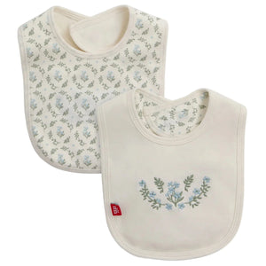 The Magnetic Me Eloise Organic Cotton Reversible Bib with Emb features two GOTS-certified bibs in cream with blue floral patterns: one with an all-over print, the other with a central floral design and a small red side label.