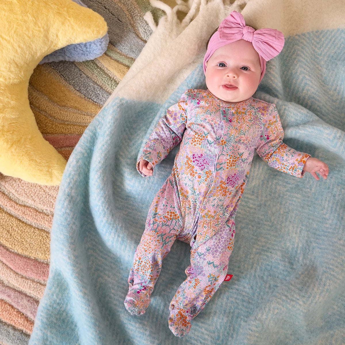 A baby in the Baby Girls’ Arabella Modal Footie by Magnetic Me with a pink bow headband lies on a blue blanket near a yellow crescent pillow.