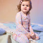 A toddler with brown curly hair and a pink bow sits on a pastel blanket, wearing the Baby Girls’ and Girls’ Arabella Modal Long Sleeve Pajama Set by Magnetic Me, looking at the camera.