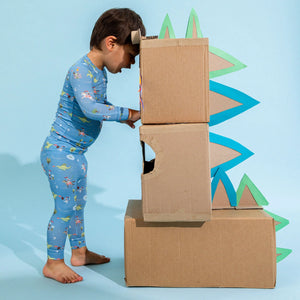 A young child wearing the Magnetic Me Baby Boys' and Boys' Good Knight Modal PJ Set stands next to large cardboard boxes decorated with green and blue dinosaur cutouts.