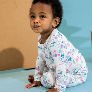 A toddler with short curly hair sits on the floor wearing Magnetic Me’s Baby Girls' and Girls' Tail of Magic Modal Magnetic PJ Set, featuring colorful floral and castle patterns. A cardboard backdrop is behind her.