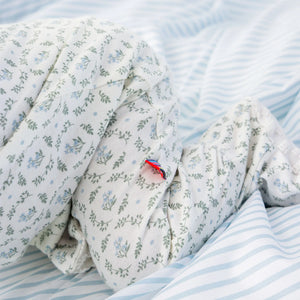 Close-up of Magnetic Me's Baby Girl's Eloise Organic Cotton Magnetic Footie, featuring GOTS certified white fabric with a blue floral print and a tiny red, blue, and white tag on a light blue and white striped background.
