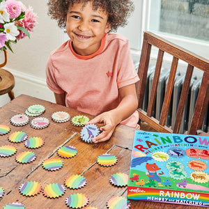 A child plays Eeboo's Rainbow Memory Matching Game at a wooden table, smiling as colorful game pieces and the box help boost concentration and color recognition skills.