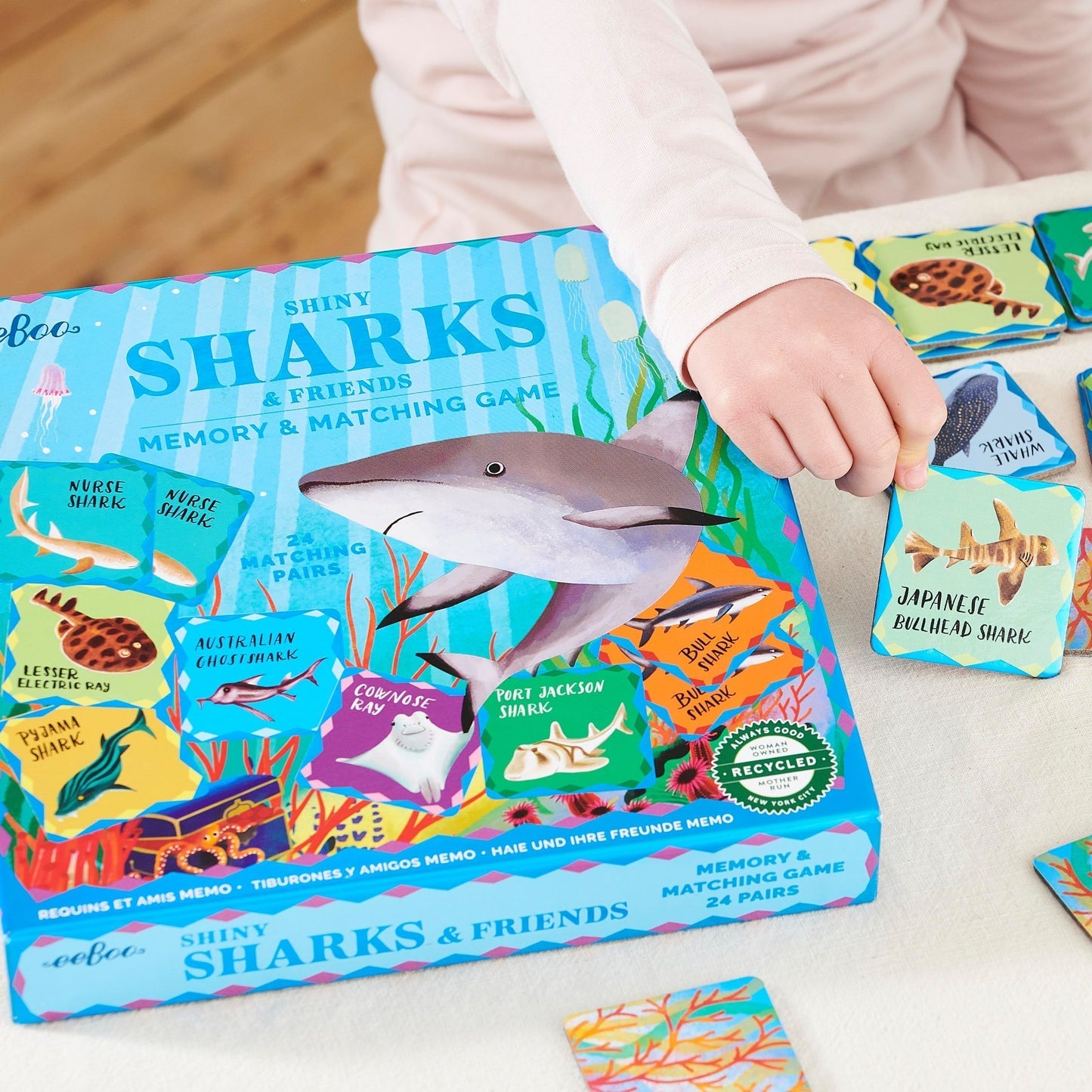 A child enjoys Eeboo's Sharks & Friends Shiny Memory Matching Game, featuring illustrated shark cards and vibrant marine life in a colorful box.