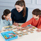 An adult and two children sit at a table playing Eeboo’s Sharks & Friends Shiny Memory Matching Game, an educational activity featuring colorful cards.