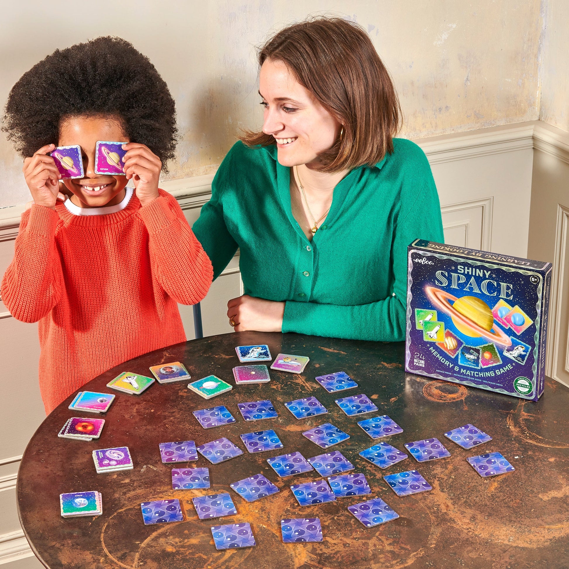 A child sits beside an adult at a table, holding two cards over their eyes while playing Eeboo’s Space Shiny Memory & Matching Game. The open game box is on the table.