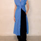 A woman wears the Callas Milano Miami Dress, a sleeveless blue stretch chambray style, over black pants and white sandals while standing against a plain beige wall.