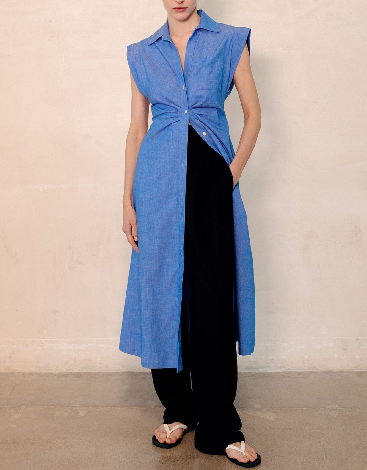A woman wears the Callas Milano Miami Dress, a sleeveless blue stretch chambray style, over black pants and white sandals while standing against a plain beige wall.