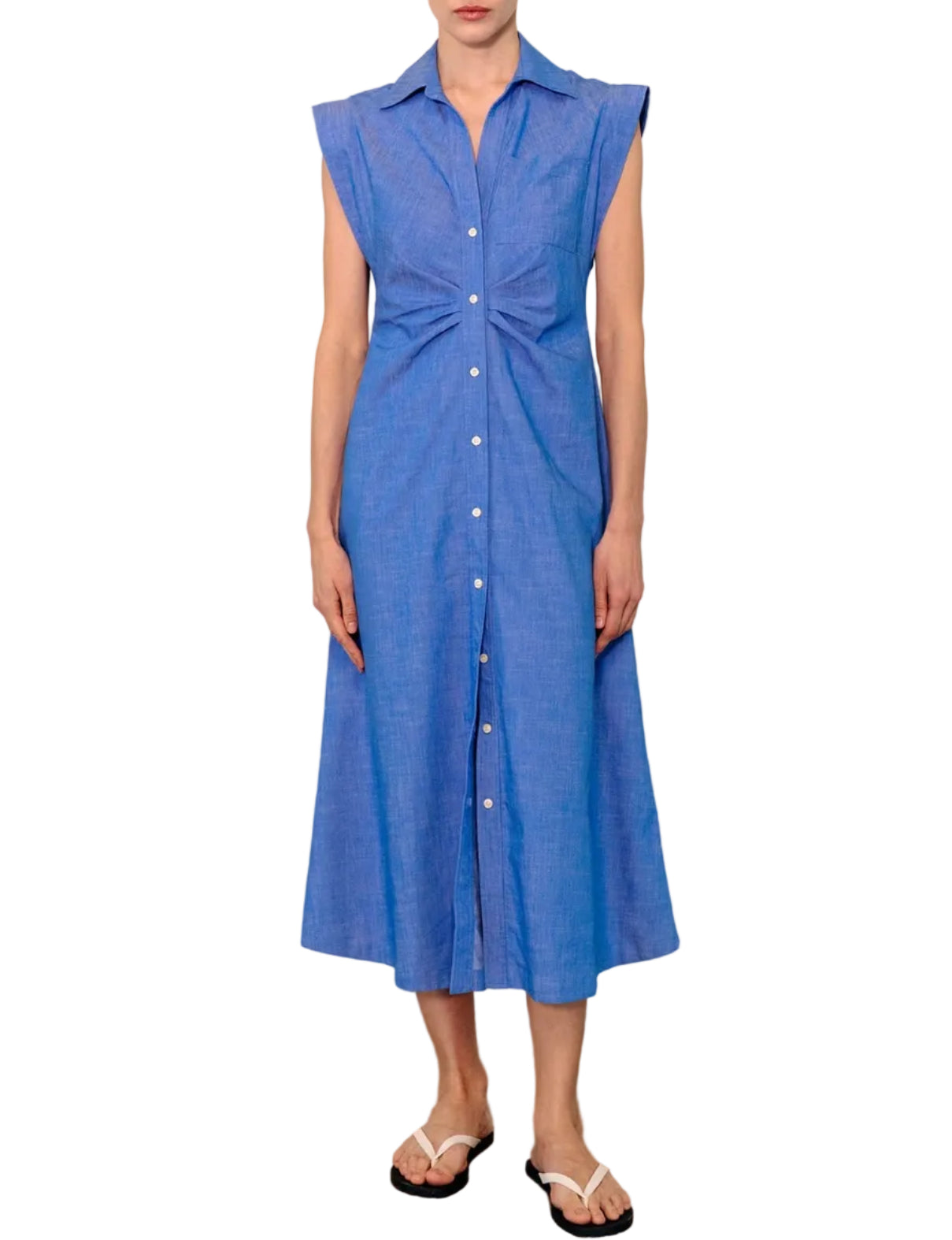 A woman stands against a white background wearing the Callas Milano Miami Dress—a sleeveless, collared, blue stretch chambray midi shirt dress—paired with flip-flops.