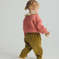A young child with curly hair faces away, dressed in a pink sweater and a floral-collared top, wearing Cozmo’s Baby Miki Wide Wale Corduroy Pants in green, and holding a small round object while standing against a plain light background.