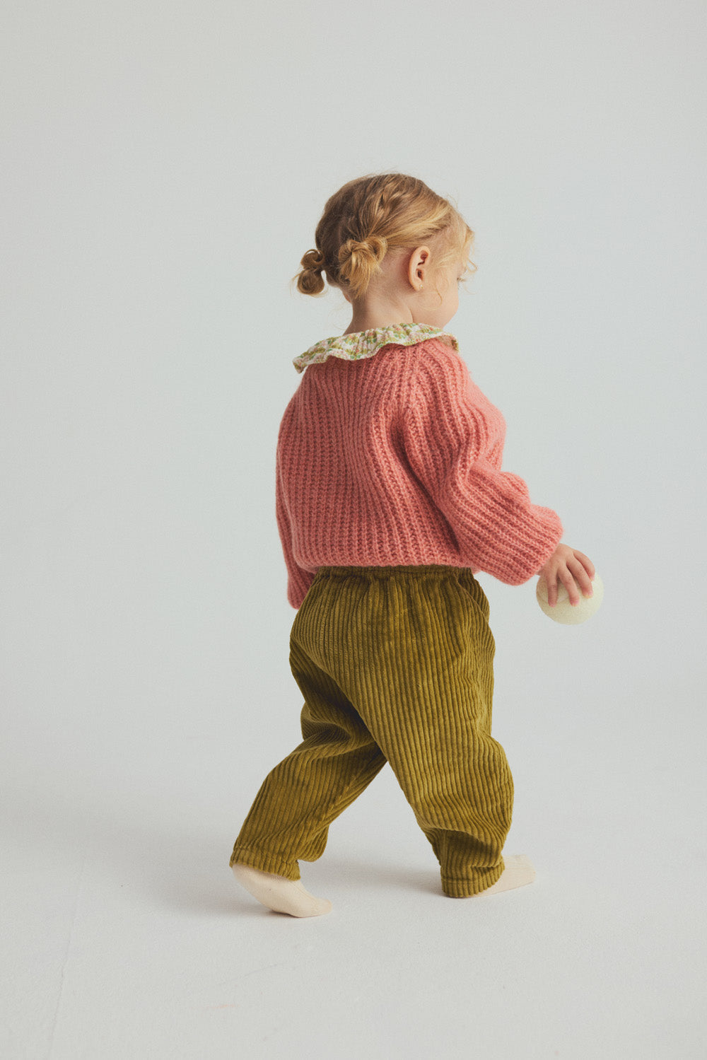 A young child with curly hair faces away, dressed in a pink sweater and a floral-collared top, wearing Cozmo’s Baby Miki Wide Wale Corduroy Pants in green, and holding a small round object while standing against a plain light background.