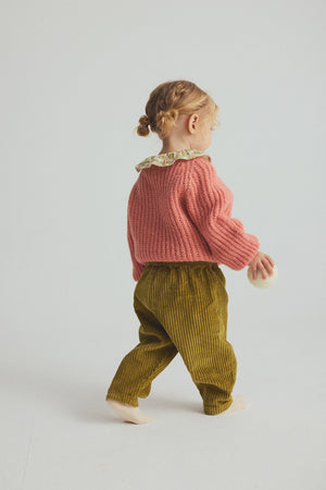 A young child with curly hair faces away, dressed in a pink sweater and a floral-collared top, wearing Cozmo’s Baby Miki Wide Wale Corduroy Pants in green, and holding a small round object while standing against a plain light background.