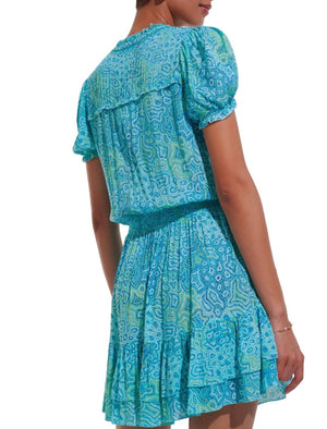 A person is wearing a blue and green Poupette St Barth Bibi Dress patterned short-sleeve dress with a smocked waist and tiered skirt. They are shown from the back.