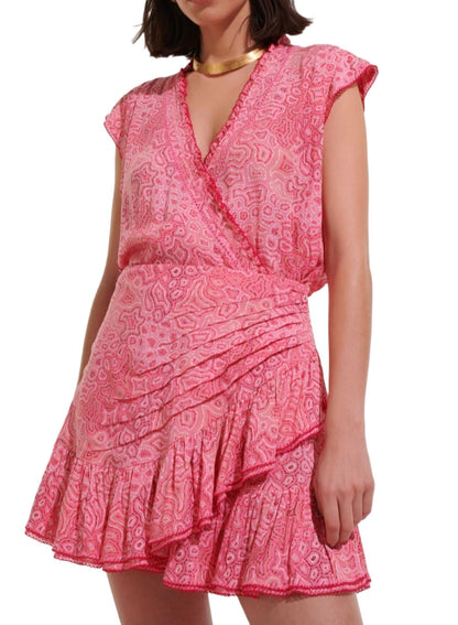 Person wearing a short, pink patterned Poupette St Barth Estelle Mini Dress with a v-neckline, cap sleeves, ruffled hem, and a gold necklace that accentuates the vibrant color.