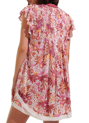 A person wearing the Poupette St Barth Sasha Mini Dress from the back, showcasing its short length and vibrant floral pattern in shades of pink, red, and orange. The beach-style dress is adorned with ruffled sleeves and a pleated upper back design.