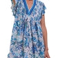 A woman wears a short, sleeveless Poupette St Barth Sasha Mini Dress adorned with blue and green floral patterns. The dress, embodying true beach style from Poupette St Barth, features a V-neckline and ruffled sleeves. Only the upper half of her body is visible.